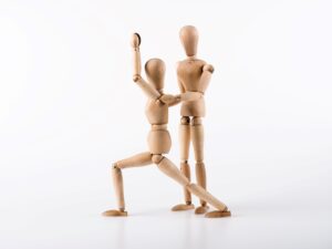 
Zwei hölzerne Gelenkpuppen demonstrieren eine Physiotherapie-Technik, wobei eine Puppe die andere in einer gestützten Pose hält. Dieses Bild symbolisiert die Bedeutung von Körpermechanik und Patientenunterstützung, Schlüsselkomponenten in den Fähigkeiten eines Physiotherapeuten, besonders relevant für Jobs in der Physiotherapie in Düsseldorf
