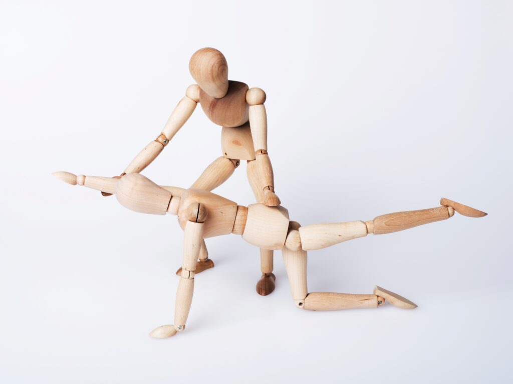 Zwei Gelenkpuppen in einer simulierten Physiotherapie-Sitzung, bei der eine Puppe eine andere manipuliert, um eine Bewegungsübung darzustellen. Die Puppen sind aus Holz und zeigen eine therapeutische Bewegung auf einem weißen Hintergrund, was die Bedeutung praktischer Fähigkeiten in Physiotherapeut-Jobs hervorhebt, speziell in einem beruflichen Kontext wie Düsseldorf